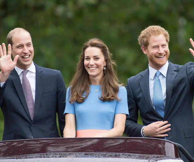 Three people in formal attire smiling and waving, standing by a car with a green, leafy background.