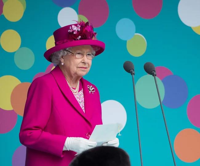 A person in a vibrant pink outfit speaks at a podium with a colorful polka dot background.