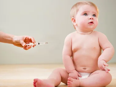 A baby sitting in a diaper looks to the side as a hand approaches with a syringe for vaccination.