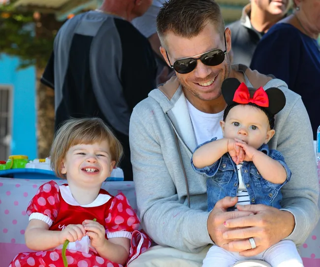 Man with sunglasses holding baby in Minnie Mouse ears, young child smiling in red polka dot dress beside him.