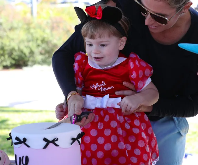 Toddler in Minnie Mouse outfit cuts birthday cake with adult's help, outdoors.