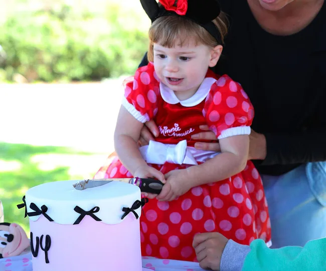 Little girl dressed as Minnie Mouse cutting a cake with help at a party.