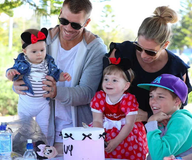 Family celebrating outdoors with two toddlers in Minnie Mouse outfits, a cake, and a boy in a purple cap.