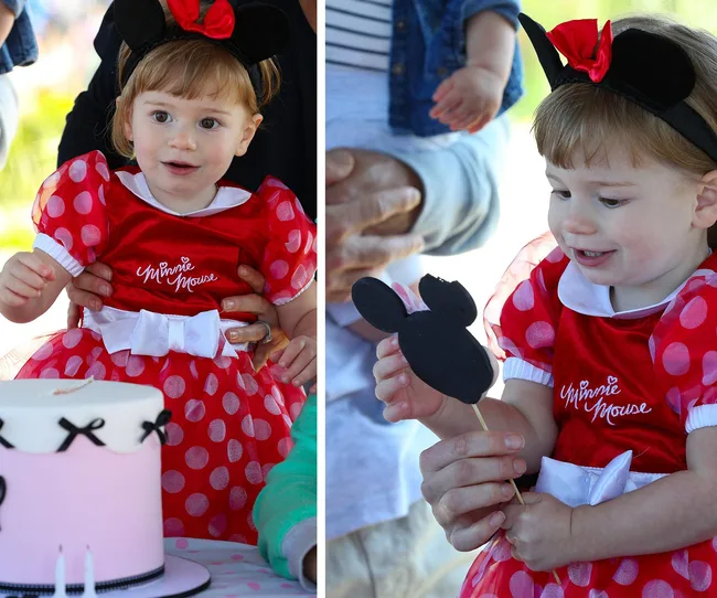 Toddler in Minnie Mouse dress holding a Mickey Mouse-shaped treat next to a cake at a party.