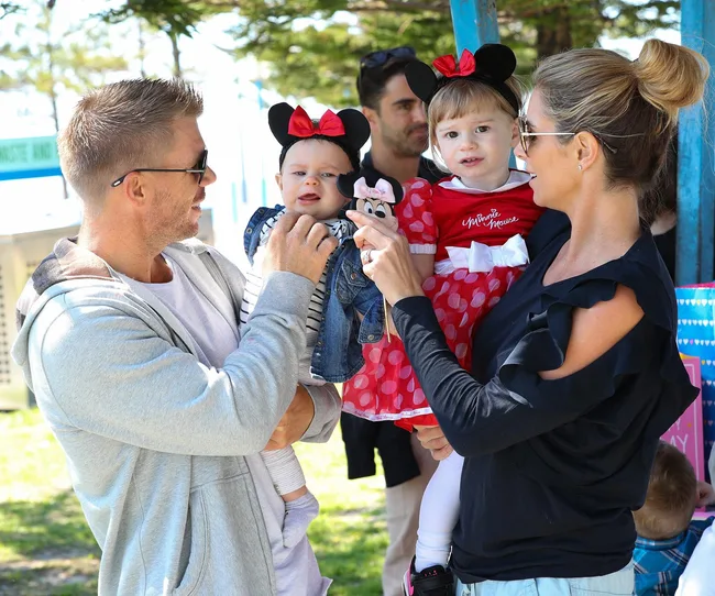 A man and woman holding two children in Minnie Mouse outfits outside.