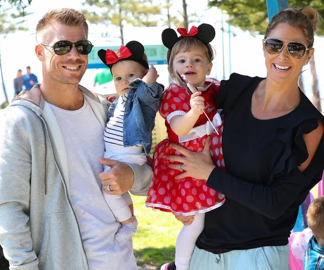 Family smiling outdoors, man holding a baby; woman with child dressed in Minnie Mouse costume.