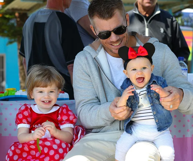 A man in sunglasses holds a smiling baby wearing a Minnie Mouse headband next to a toddler in a red polka dot dress.