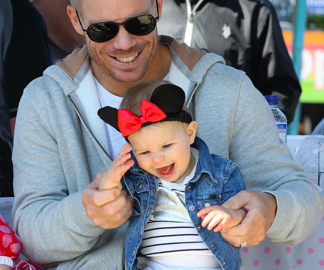 A man in sunglasses smiles while holding a baby wearing Mickey Mouse ears with a red bow, both appear happy outdoors.