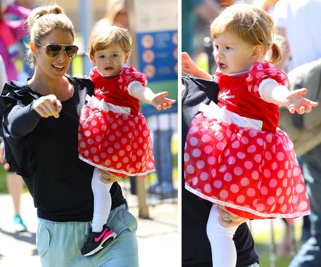 Woman holding a child in a Minnie Mouse dress outdoors; child appears curious, wearing white leggings and black shoes.