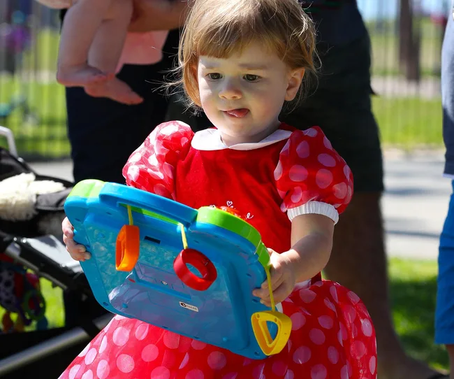 Toddler in a polka dot dress holds a colorful toy tablet outdoors.
