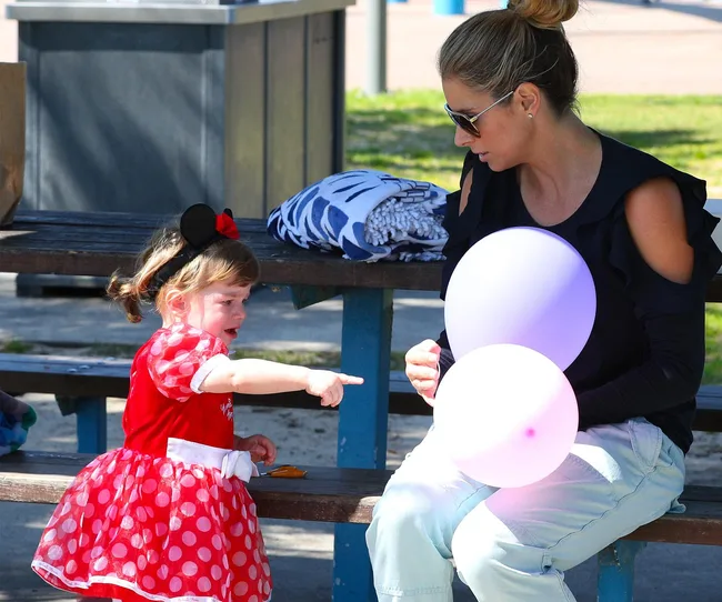 A girl in a red polka-dot dress points at a woman holding balloons at a picnic table.