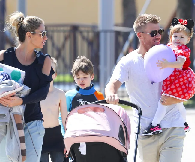 Family walking outdoors; man holding a child in red polka dot dress with a balloon, woman carrying towels.