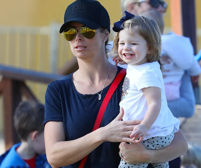 Woman in sunglasses holding a laughing toddler with a bow in her hair.