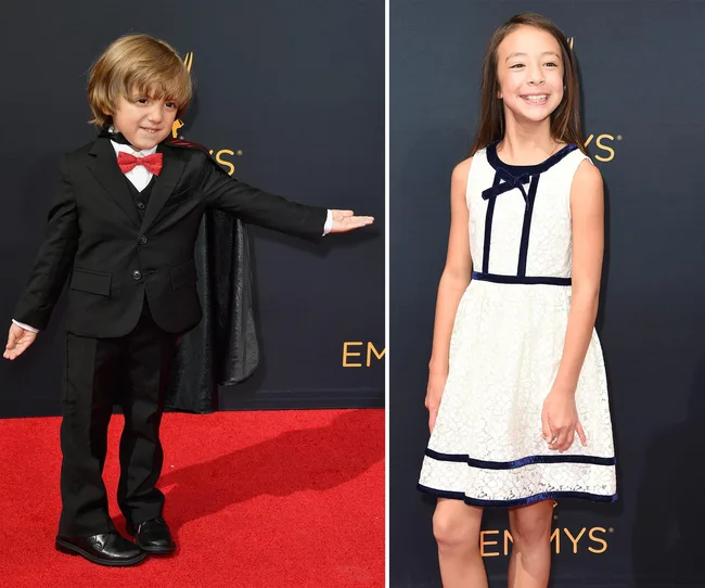 Children in formal attire on the Emmys red carpet, boy in a suit with bowtie and girl in a white dress, smiling.