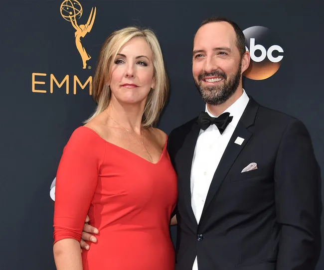 A woman in a red dress and a man in a tuxedo smiling at an Emmy Awards event backdrop.