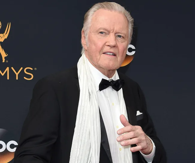 Elderly man in a tuxedo and white scarf at a formal event, standing against an Emmy Awards backdrop.