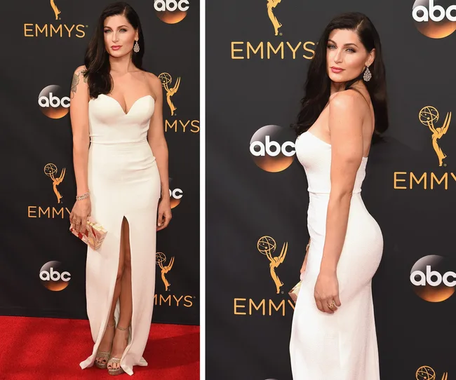 Woman in a white gown with a thigh-high slit on the Emmys red carpet, holding a clutch, with long dark hair.