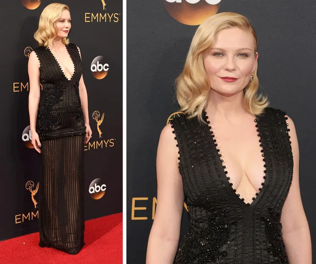 Blonde woman in a deep V-neck black gown on a red carpet at the Emmys, with a logo in the background.