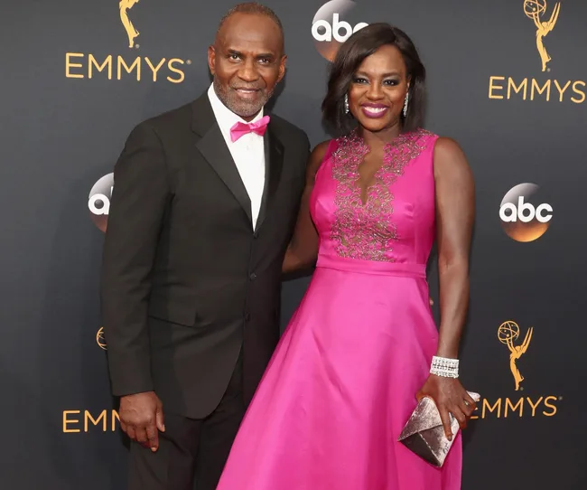 Couple posing on the Emmy Awards red carpet, woman in pink dress, man in suit, smiling.