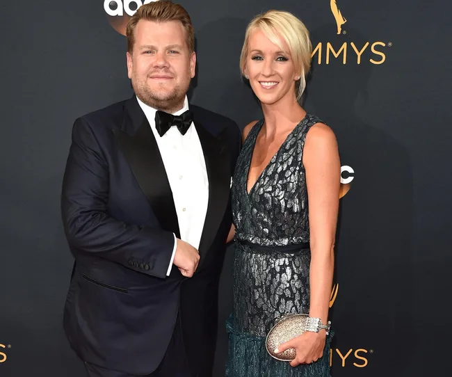 A person in a suit and bow tie poses with a person in a sleeveless dress on a red carpet event with "Emmys" in the background.