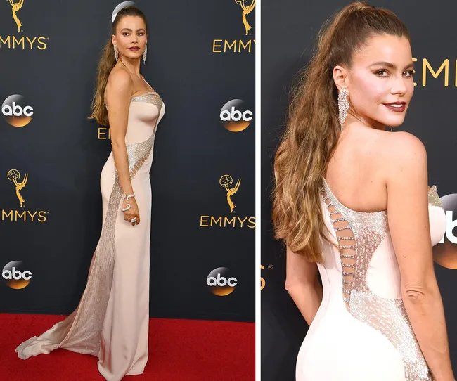 Woman in an elegant gown poses on the red carpet at the Emmys, with backdrop featuring logos.