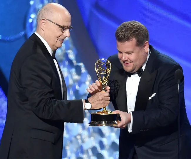 Two men in tuxedos on stage, one handing an Emmy Award to the other.