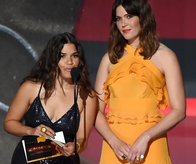 Two women standing on stage, one in a dark sequin dress holding a microphone, the other in a yellow ruffled dress.