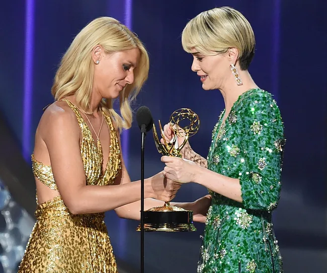 Two women on stage exchanging an Emmy award, one in a gold dress and the other in a green dress.