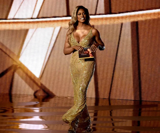 Elegant person in a shimmering gold dress on stage, holding a card at an awards ceremony.