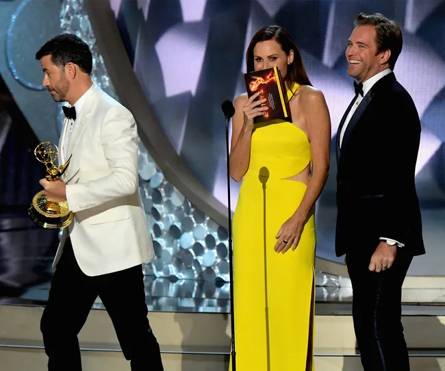 Three people at an awards show; one holds a trophy, one in yellow speaks into a card, and the other smiles.