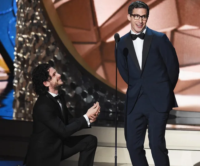 A man on stage kneels playfully as if proposing to another man during an award show. Both are in suits.