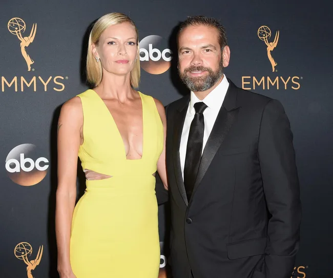 A man in a suit and a woman in a yellow dress pose at an Emmy Awards event backdrop.