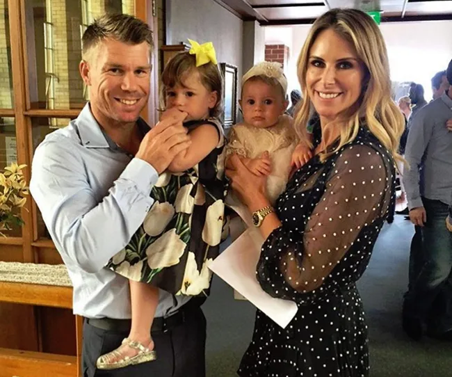 A smiling couple holding two young children, one with a yellow bow, in a warmly lit indoor setting.
