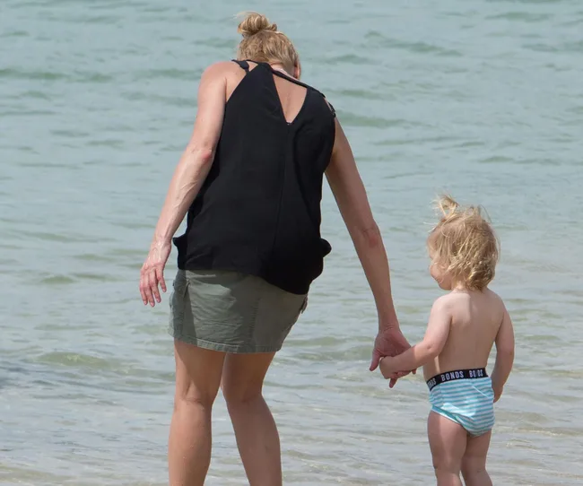 Adult and toddler holding hands, walking in shallow beach water, with the adult wearing a black top and khaki shorts.