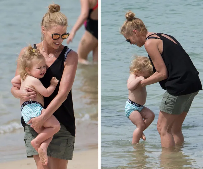 A woman in sunglasses holds a toddler at the beach, both near the water's edge.