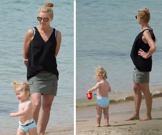 A woman in sunglasses and a black top with a child in swimwear playing on the beach near the water.