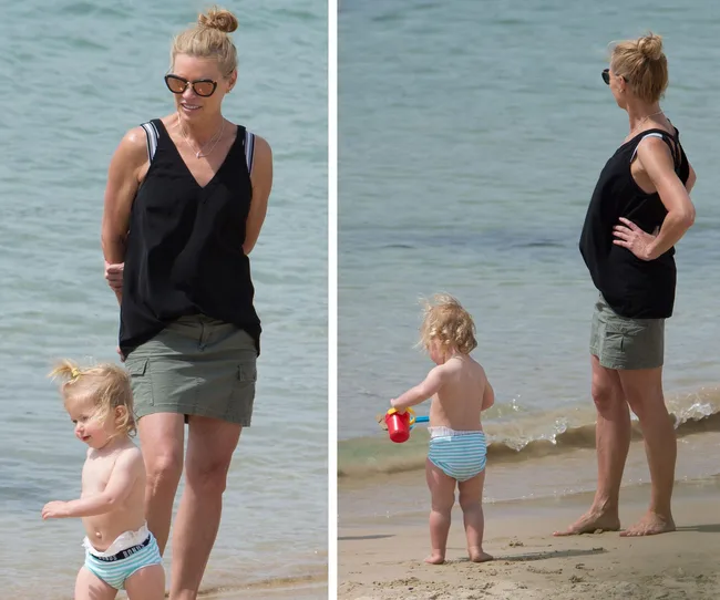 Woman and toddler on a beach, woman in black top and shorts, toddler in striped swimsuit, playing near the water.