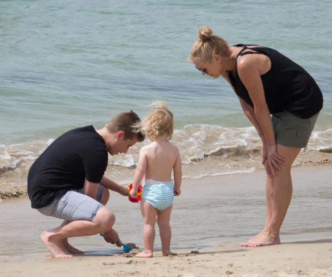 Three people playing by the ocean: an adult crouches, a child in a diaper stands, and a woman looks on, smiling.