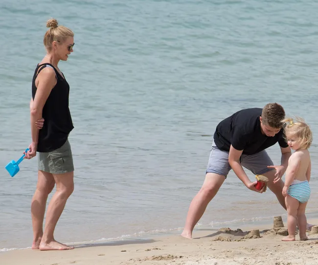 A woman, boy, and young child build sandcastles on a beach. The woman holds a blue shovel.