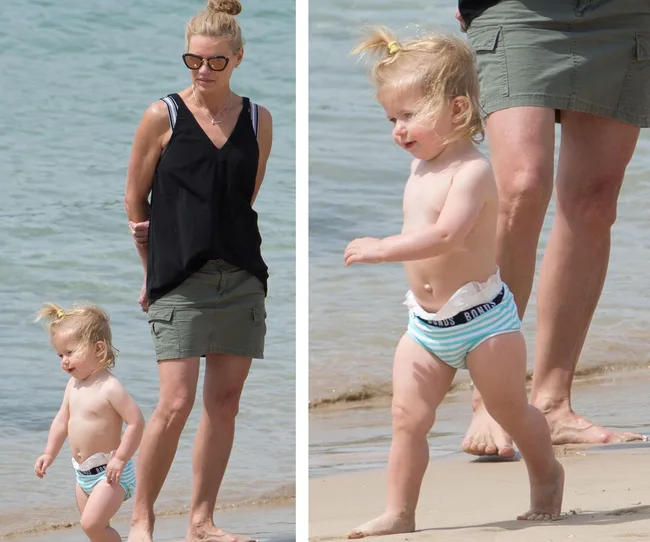 Woman in sunglasses and child in striped shorts walking on the beach near the water.