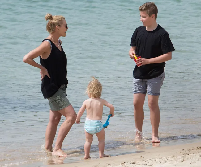 A woman and two kids playing on the beach by the water, with a toddler holding a blue shovel.