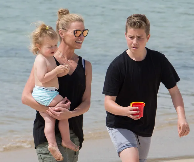 A woman with sunglasses holds a toddler, while a boy with a red cup walks on a beach.