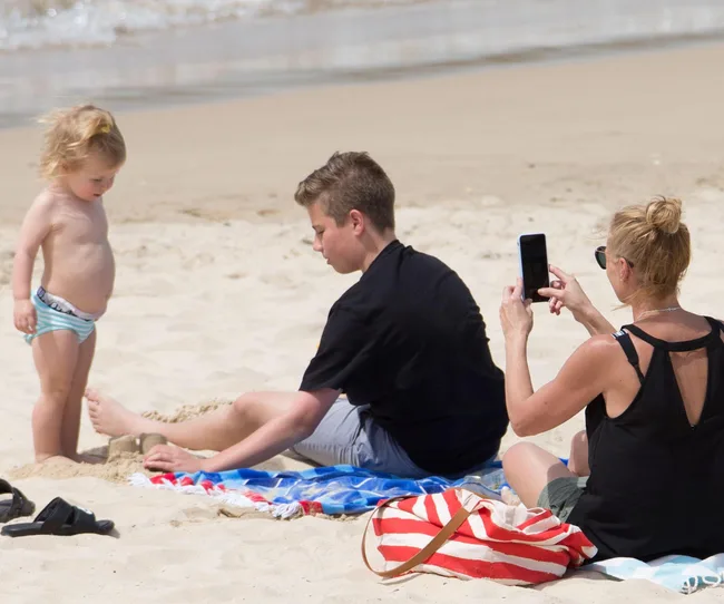 Family on beach, child in swimwear stands, teenager sands, woman takes phone photo. Beach towel and bag nearby.