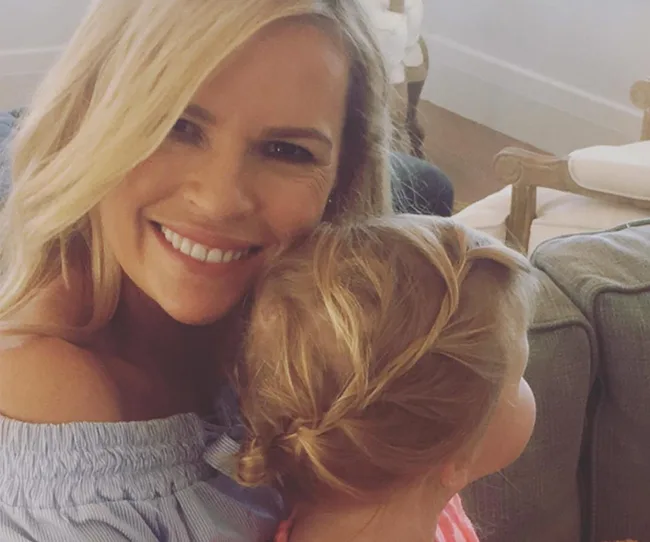 Blonde woman smiling while hugging a child with braided hair, in a cozy living room setting.