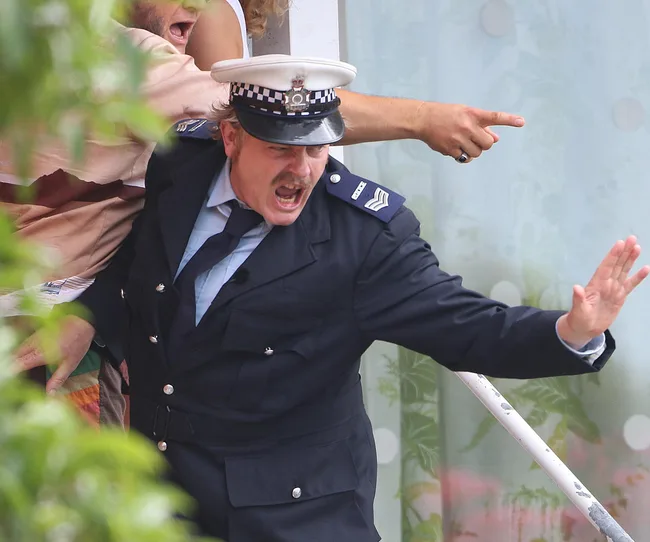 Police officer in uniform shouting with hand raised, another person pointing in the background.
