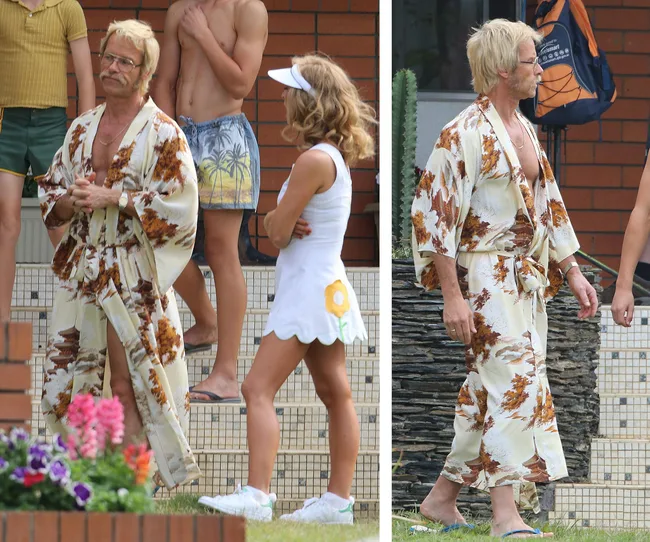 Actor on set in floral robe talking to woman; 'The Greatest Showman' theme.