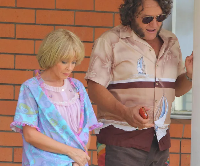 A woman in a colorful robe and a man in a patterned shirt stand against a brick wall, the man holding a phone.