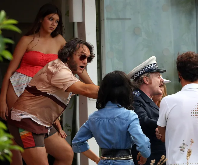 A group of people discuss animatedly outside a building; one man is dressed as a police officer.
