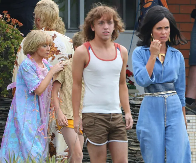 Group of people in 70s-style clothing, including a person in a blue jumpsuit, outdoors near a brick building.