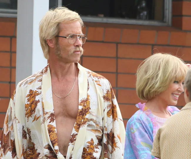 A man in 70s style clothes with glasses and a mustache stands next to a woman with short blonde hair, both in colorful outfits.